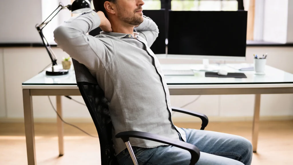 Office worker leaning back in his chair with hands behind his head, stretching his chest and shoulders during a workday reset break.