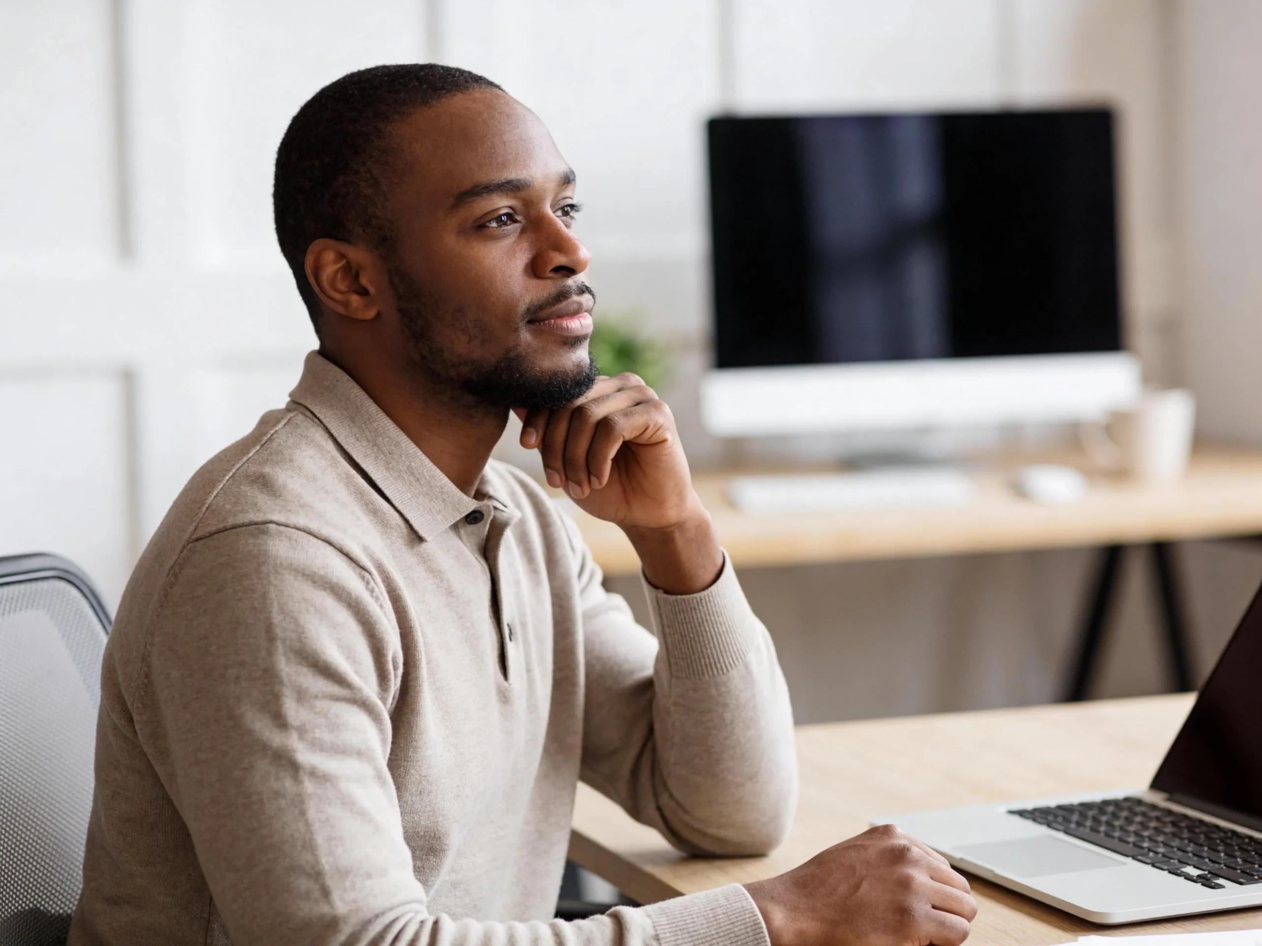 Man sitting at a desk in a modern office, pausing thoughtfully with his hand resting on his chin during a short workday reset break.